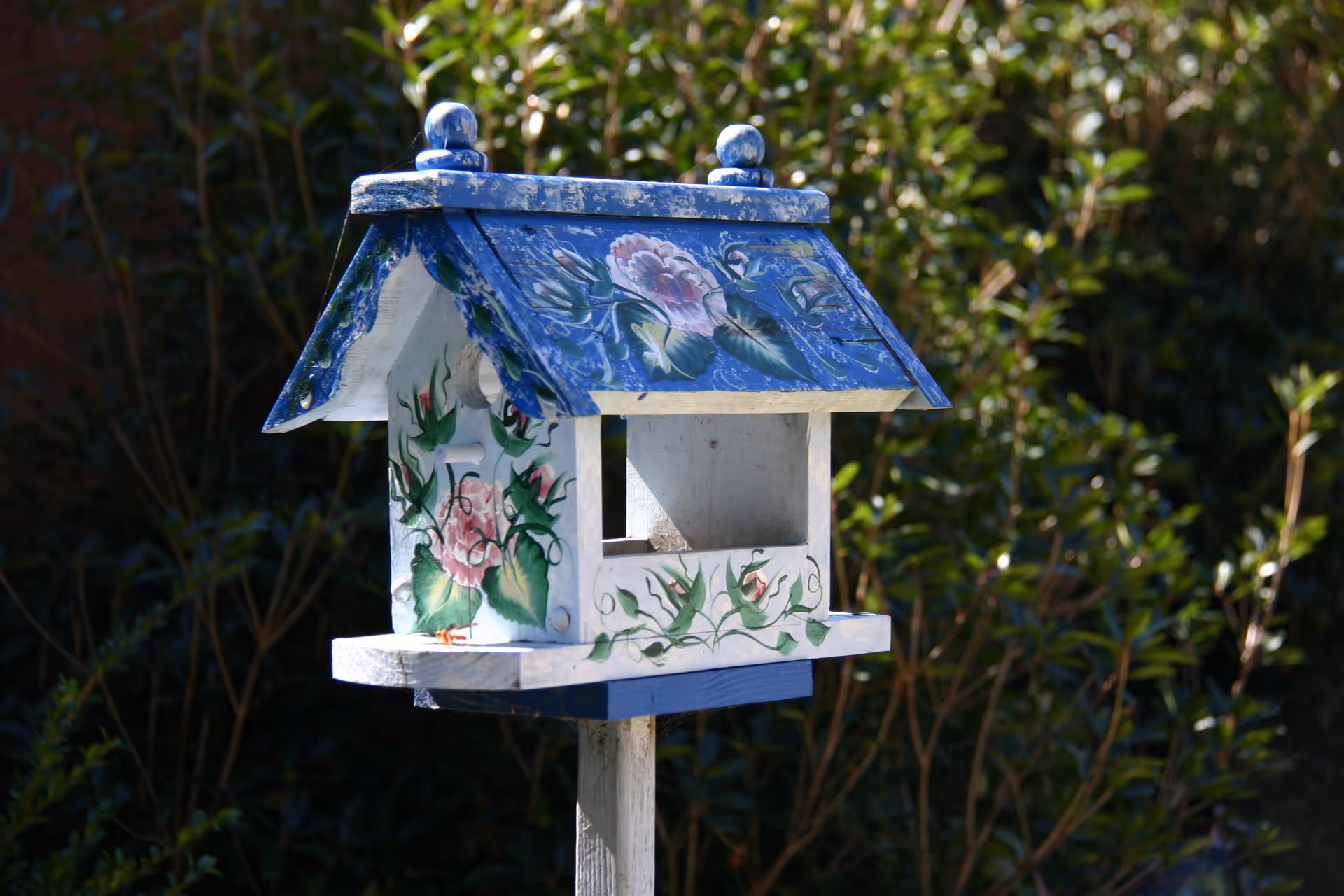 A decorative birdhouse painted with blue roof and floral designs, mounted on a wooden post with green foliage in the background.