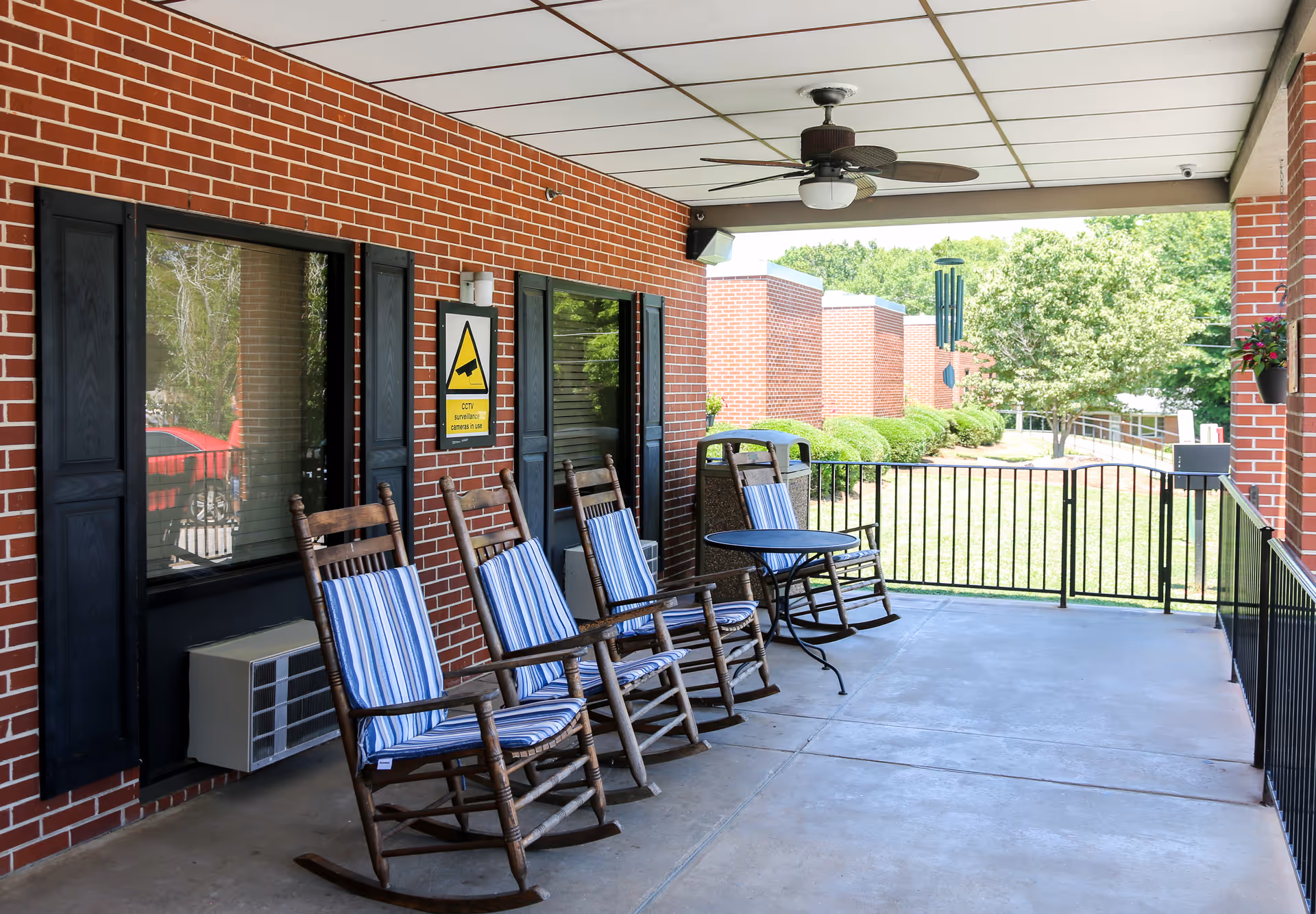 Covered outdoor patio area with four wooden rocking chairs with blue and white striped cushions, a small round black table, a ceiling fan, and a brick wall with windows and a CCTV surveillance sign. The patio overlooks a grassy area with bushes and trees.