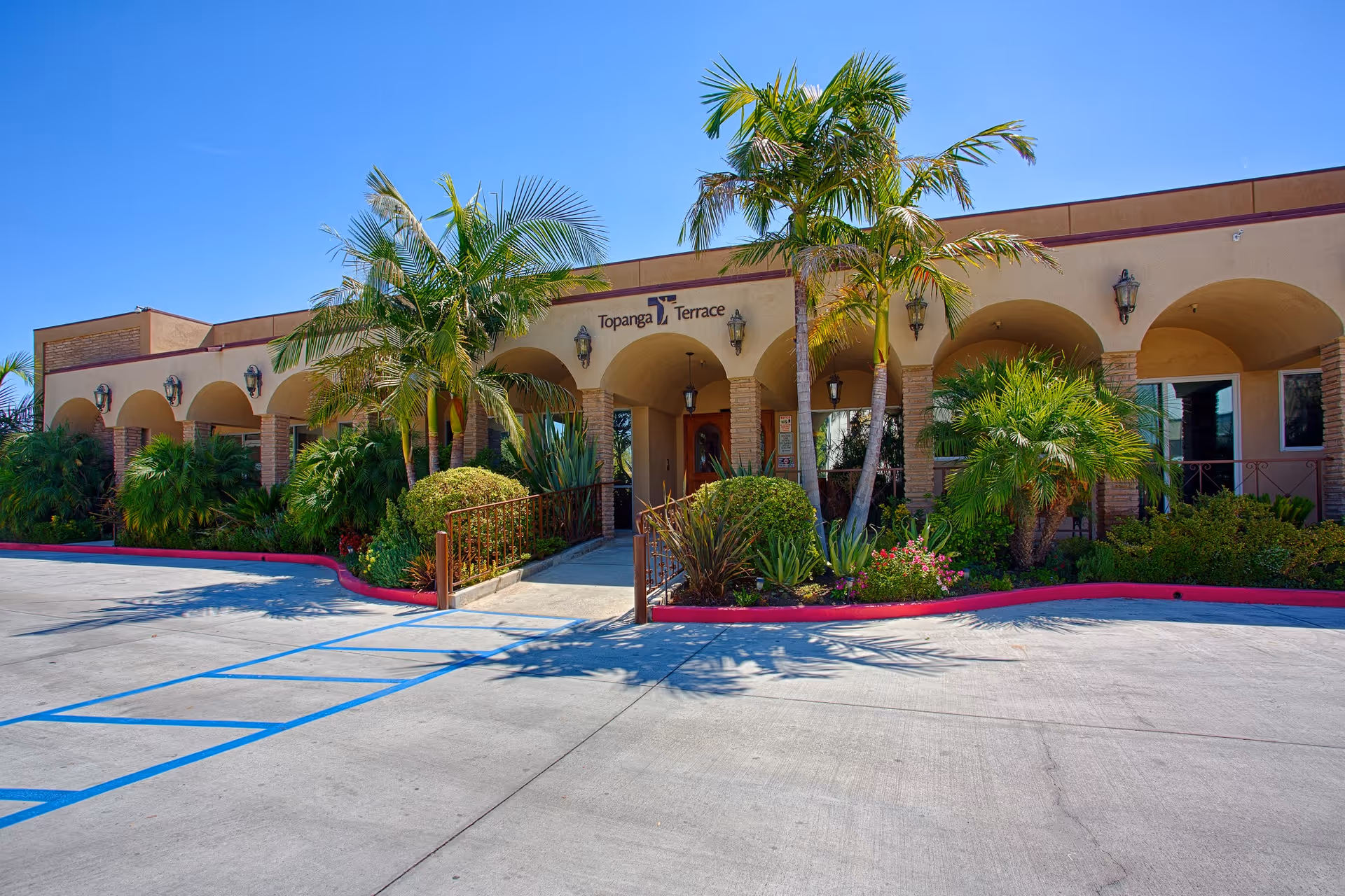 Exterior view of Topanga Terrace Rehabilitation and Subacute building with arched entryway, palm trees, and landscaped greenery under a clear blue sky. The parking area in front has marked handicap spaces.