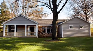 Exterior view of a single-story residential building with a covered porch, multiple windows, and a large tree in the front yard with green grass.