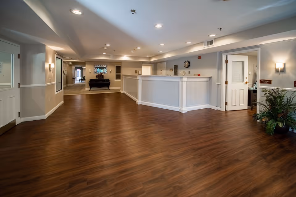 Interior view of a senior living facility reception area with wooden flooring, a white reception desk, beige walls, recessed ceiling lights, wall sconces, a clock on the wall, a potted plant, and an open door leading to an office labeled 'Hospitality Suite'.