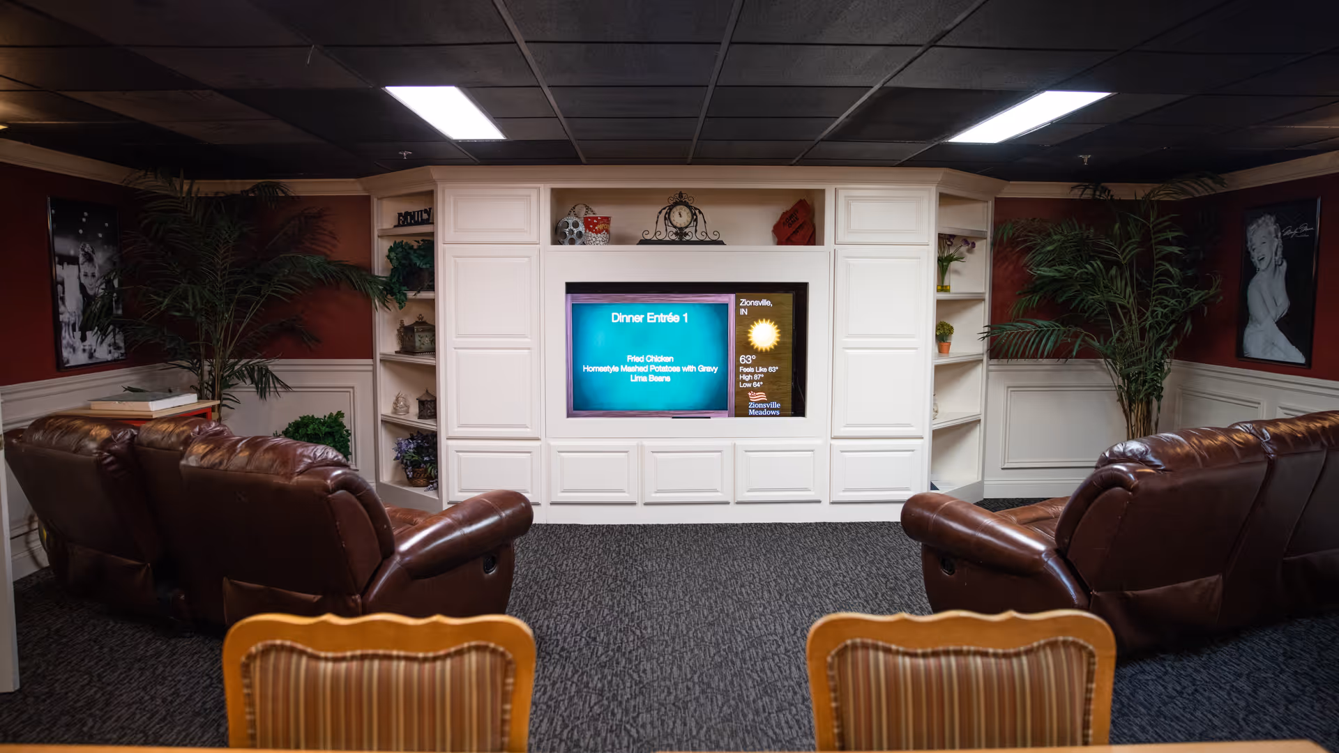 A cozy lounge area with two brown leather recliners facing a built-in white entertainment center. The entertainment center has a TV displaying dinner menu and weather information for Zionsville Meadows. The room features carpeted flooring, potted plants on either side of the entertainment center, and framed black and white photos on the walls.