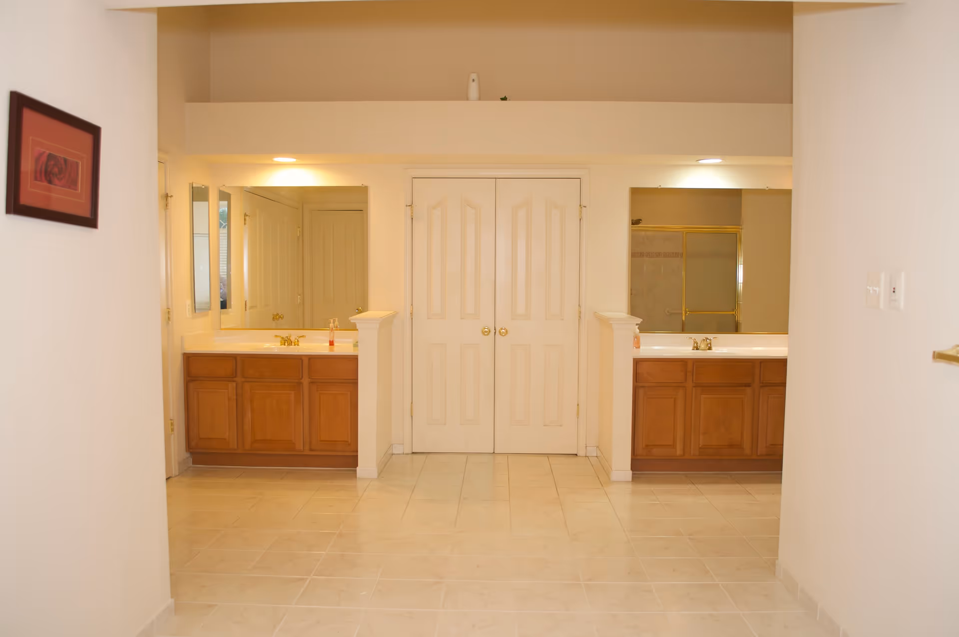 A spacious bathroom area with two separate wooden vanities, each with a sink, mirror, and overhead lighting. Between the vanities is a set of double doors. The floor is tiled in a light color, and there is a framed picture on the left wall.