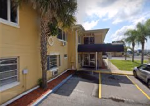 Front exterior of The Villages Senior Living with a yellow building, walkway, and palm trees.