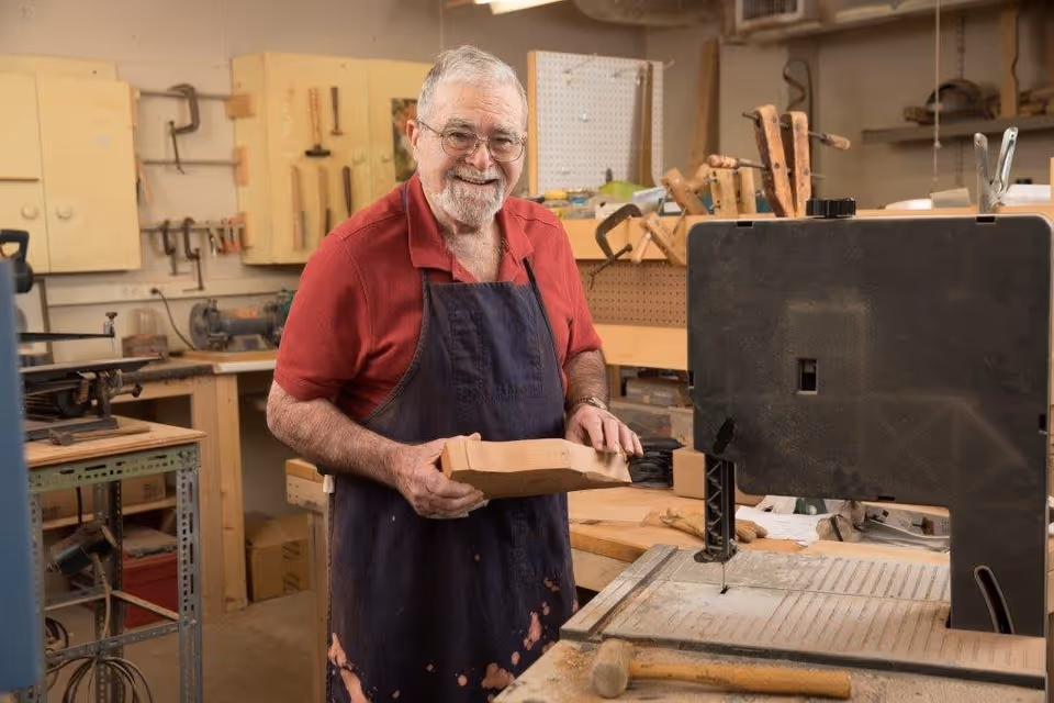An elderly man wearing glasses, a red polo shirt, and a dark apron stands in a woodworking workshop holding a piece of wood. The workshop is equipped with various woodworking tools and machines, including a bandsaw in the foreground and clamps on the wall behind him. The man is smiling and looking at the camera.