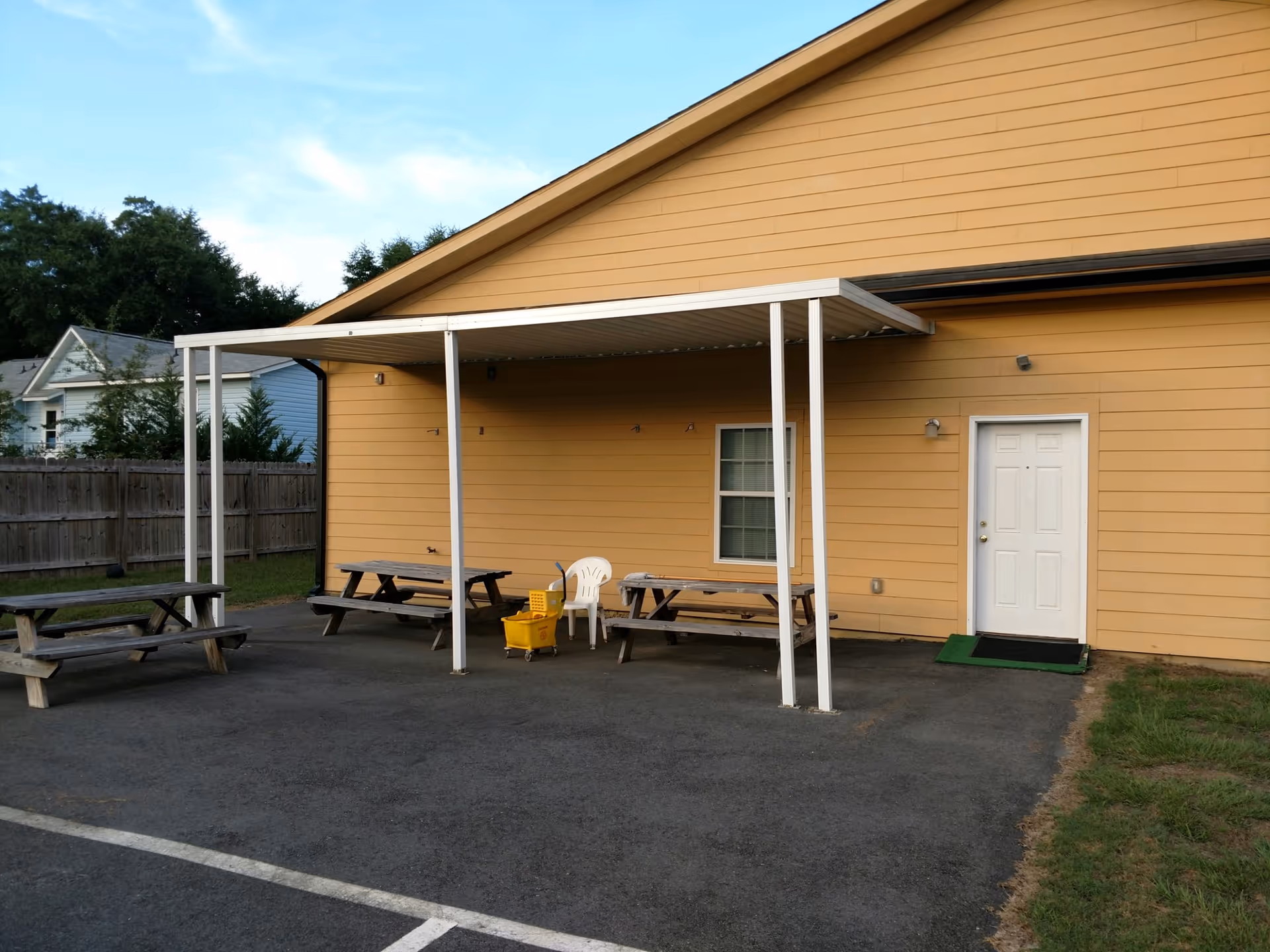 Outdoor area of a facility with a yellow building wall, a white door, a window, and a white metal canopy covering two wooden picnic tables and a white plastic chair. There is a yellow mop bucket under the canopy and a wooden fence and trees in the background.