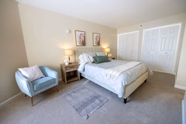 A bright and clean bedroom with a large bed featuring a tufted headboard, white and light blue bedding, and decorative pillows. On either side of the bed are wooden nightstands with lamps and small decor items. A light blue armchair with a white pillow is positioned to the left of the bed. The room has beige walls, a light carpeted floor, two framed floral artworks above the bed, and white double-door closets on the far wall.