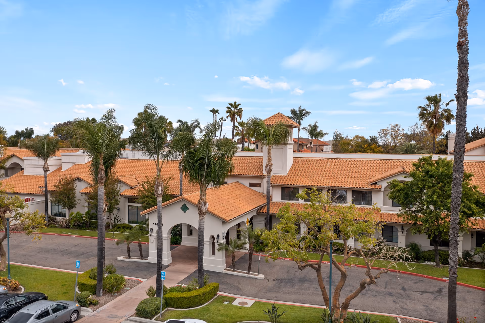 Exterior view of Oakmont of Chino Hills senior living facility showing a building with terracotta roof tiles, white walls, palm trees, and a covered entrance driveway under a blue sky with scattered clouds.
