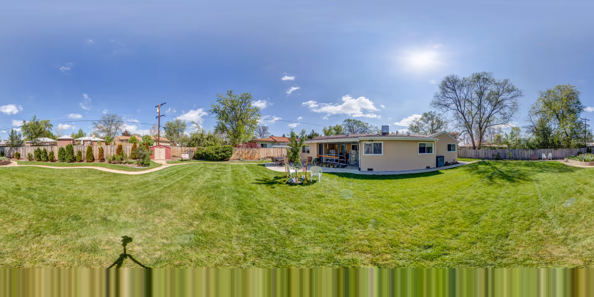A wide panoramic view of a large, well-maintained grassy backyard area at Jaxpointe Assisted Living at Flower Ct Memory Care Home. The yard is surrounded by a wooden fence with trees and shrubs along the perimeter. There is a single-story building with a covered porch on the right side, and a small garden area with chairs in the center. The sky is clear with a few clouds and the sun is shining brightly.