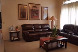 A living room with two brown leather sofas, a coffee table topped with a plant and runner, framed artwork on the wall, and a side table with a lamp by a curtained window.