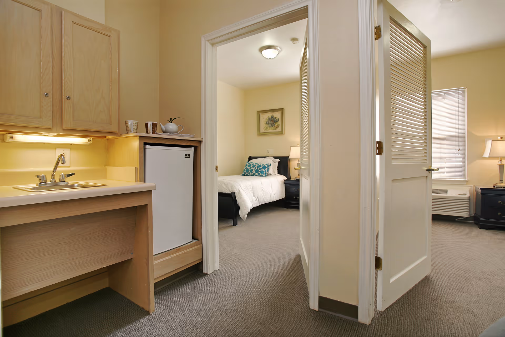 Interior view of a senior living facility room showing a small kitchenette with a sink, cabinets, and a mini refrigerator on the left. Two open doors lead to a bedroom with a bed, nightstand, lamp, and framed artwork on the wall. The room has beige walls and carpeted floors.