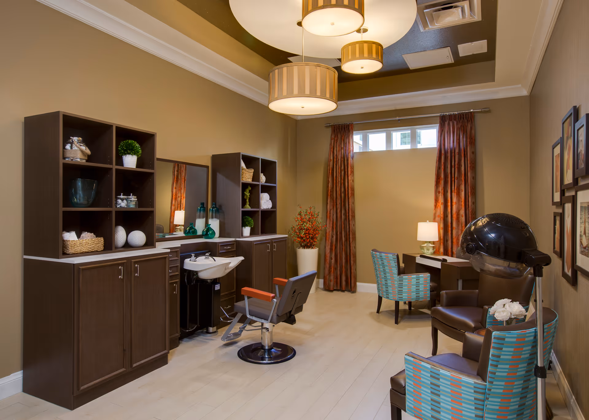 Interior of a salon room in a senior living facility with a hair washing station, salon chair, shelves with decorative items, two patterned chairs, a hair dryer chair, a desk with a lamp, and framed pictures on the wall.