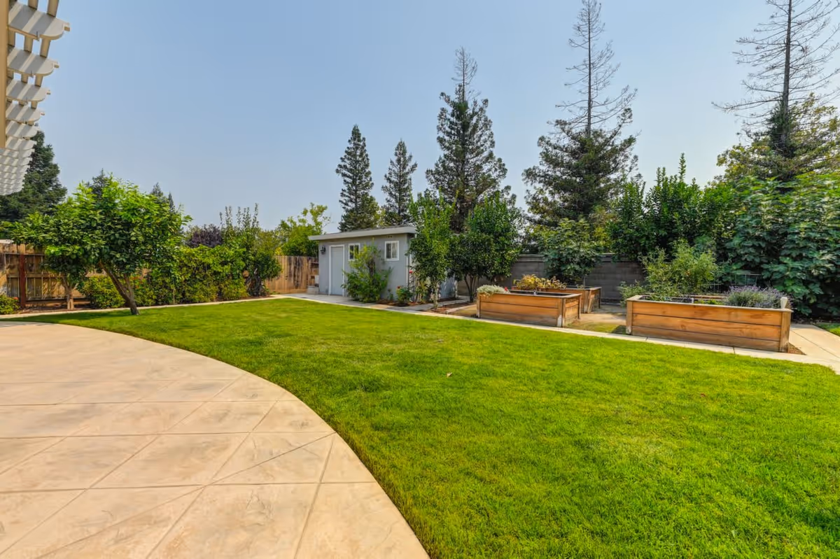A well-maintained outdoor garden area with a green lawn, raised wooden garden beds, various trees and shrubs, a small shed, and a paved patio area under a pergola.