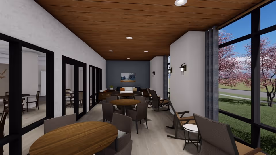 A bright and modern common area with round wooden tables and chairs, rocking chairs near large windows with views of green grass and pink blossoming trees. The room has a wooden ceiling with recessed lighting, white walls with sconces, and a blue accent wall with a landscape painting. Adjacent rooms with more seating are visible through glass doors on the left.