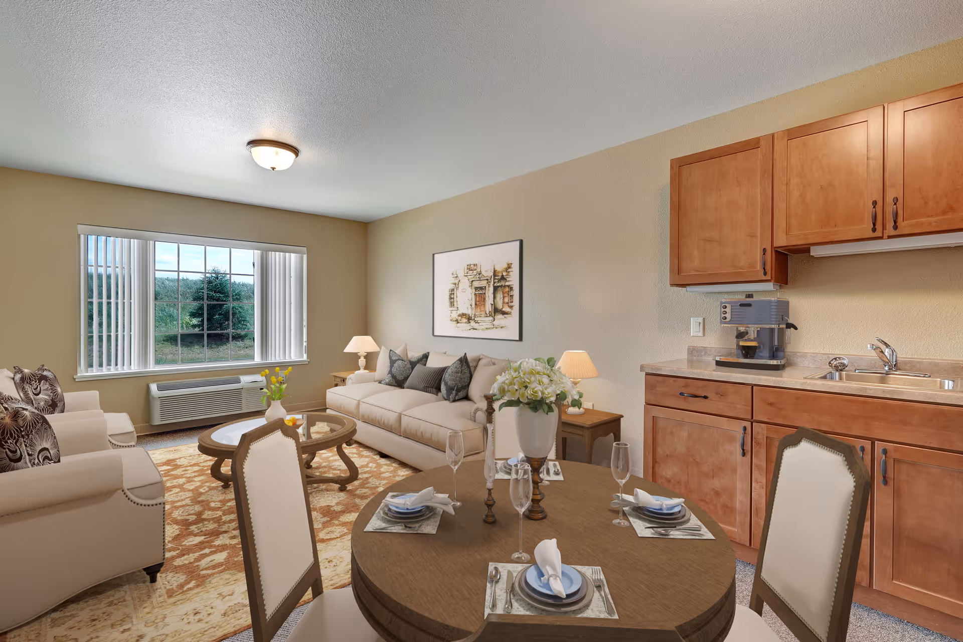 A cozy living area in a senior living facility featuring a beige sofa with decorative pillows, two armchairs, a round wooden dining table set with plates, napkins, and glasses, a coffee table with a vase of flowers, wooden cabinetry with a sink and coffee machine, and a large window with vertical blinds letting in natural light.