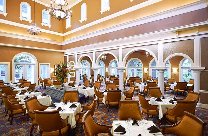 Spacious elegant dining room with high ceilings, arched columns, chandeliers, and multiple tables set with white tablecloths and place settings.