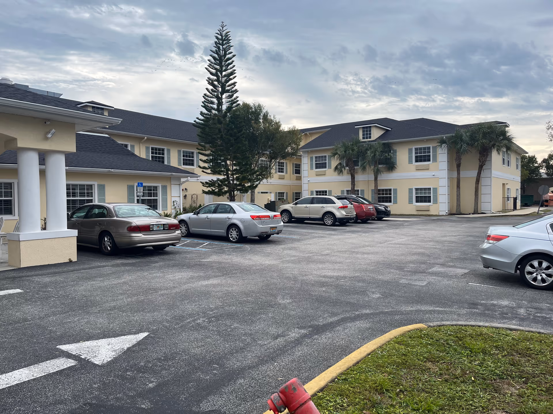 Parking lot in front of a two-story beige assisted living facility building with several parked cars, palm trees, and a cloudy sky.