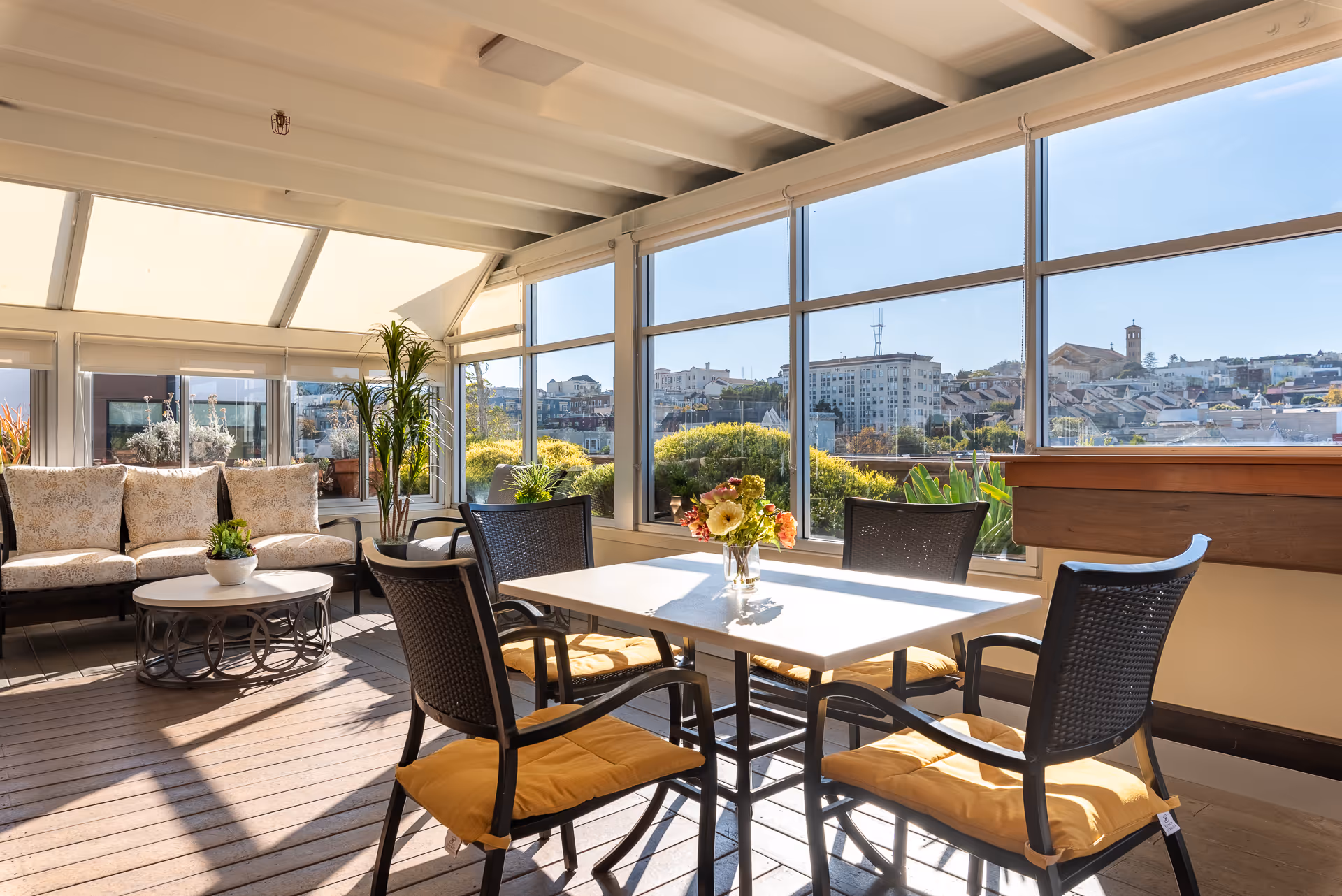 Sunlit indoor seating area with a dining table and chairs, a cushioned sofa, potted plants, and large windows showing a city view.