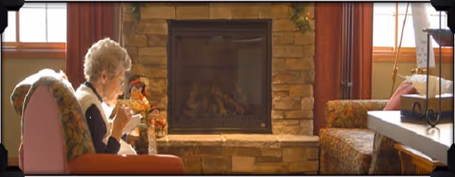 An elderly woman sits in a cushioned chair sipping a beverage in a cozy common room beside a stone fireplace.