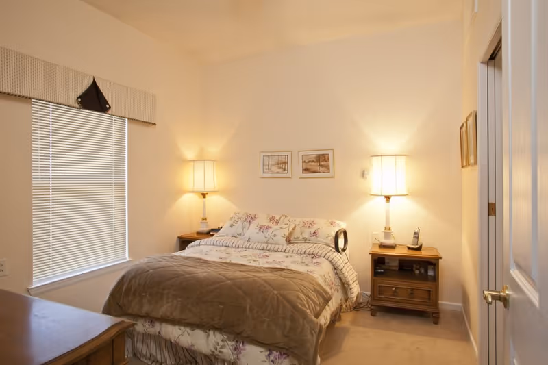 A cozy bedroom with a double bed covered in floral and striped bedding, flanked by two wooden nightstands each with a lamp. There are two framed pictures on the wall above the bed, a window with blinds and a valance on the left, and a door partially open on the right.