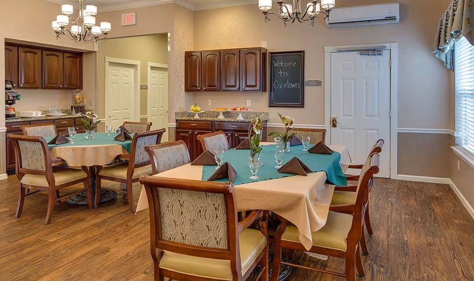 Dining room with two tables covered with beige and teal tablecloths, each set with glasses, napkins, and small flower arrangements. Wooden chairs with patterned upholstery surround the tables. The room has wooden flooring, beige walls, wooden cabinets, and a chalkboard sign that says 'Welcome to Capetown'. There are chandeliers hanging from the ceiling and a window with curtains on the right side.