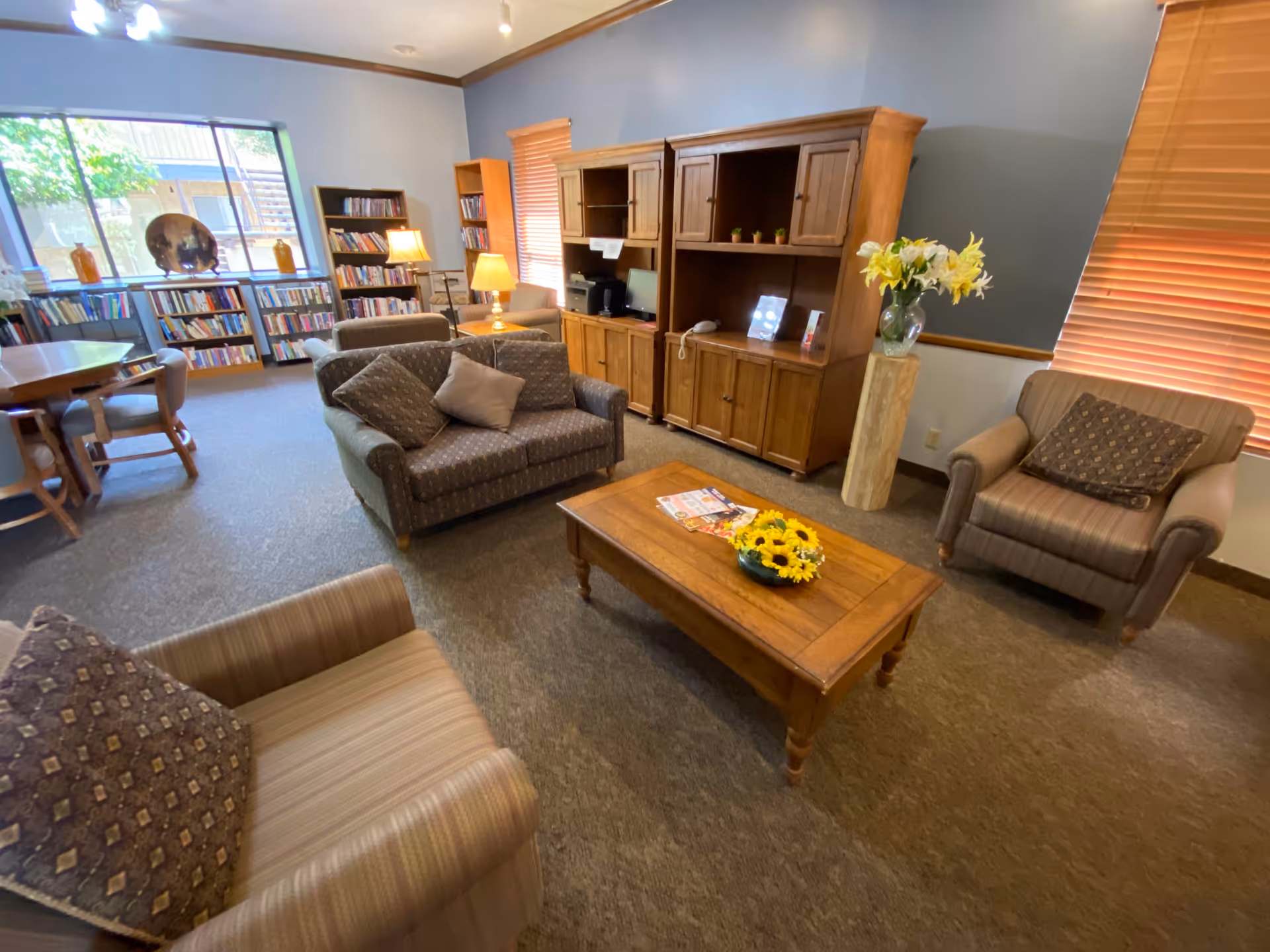 A cozy senior living common area with comfortable armchairs and a loveseat arranged around a wooden coffee table with a bowl of sunflowers and magazines. There is a wooden entertainment center with shelves and cabinets, a computer, and a telephone. Bookshelves filled with books line the walls near large windows letting in natural light. A round wooden table with chairs is visible in the background, along with lamps providing warm lighting.