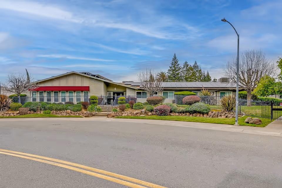 Exterior front view of a single-story assisted living facility building with a red awning, surrounded by well-maintained landscaping including bushes, trees, and a rock border. There is a sidewalk and a street with a double yellow line in the foreground, and a streetlamp on the right side.