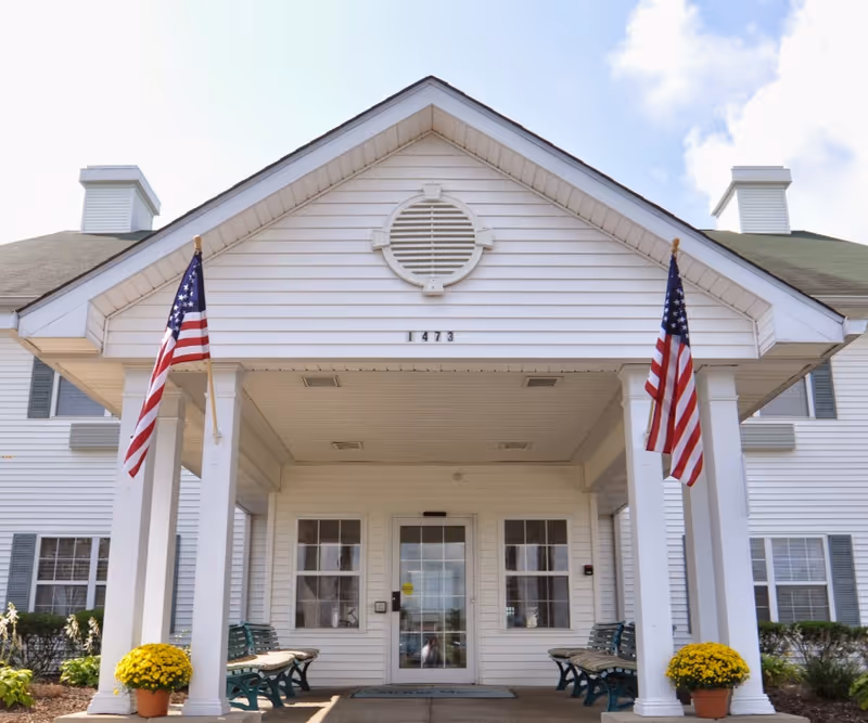 Front view of a white senior living facility entrance with flags and benches.