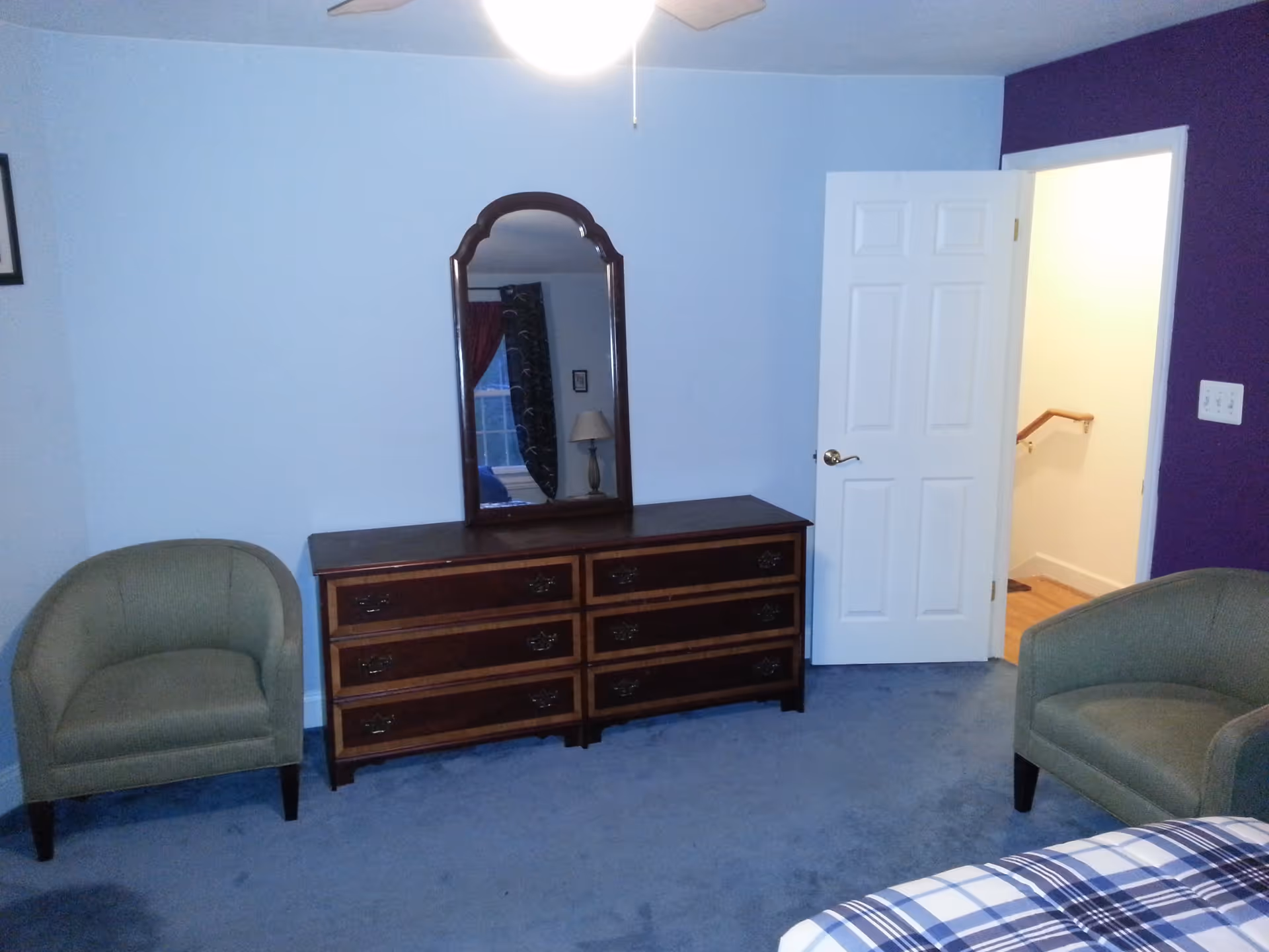 A bedroom with light blue walls and a purple accent wall. The room features a wooden dresser with six drawers and a large mirror on top. Two green upholstered armchairs are placed on either side of the dresser. A white door is open, revealing a hallway with wooden flooring and a handrail. Part of a bed with a blue and white plaid blanket is visible in the foreground.