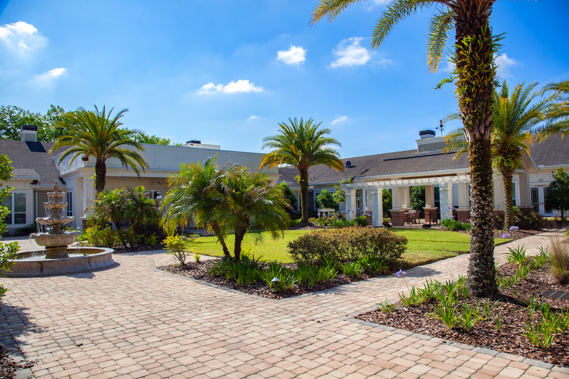 Outdoor courtyard area of a senior living facility with palm trees, a multi-tiered stone fountain, paved walkways, green grass, and a covered patio with seating under a clear blue sky.
