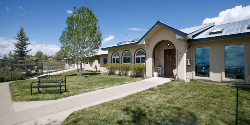 Exterior view of a single-story building with a metal roof and arched windows. There is a concrete walkway leading to a front door decorated with a wreath. The building is surrounded by a well-maintained lawn, a black metal bench, a birdbath, and several trees under a partly cloudy blue sky.