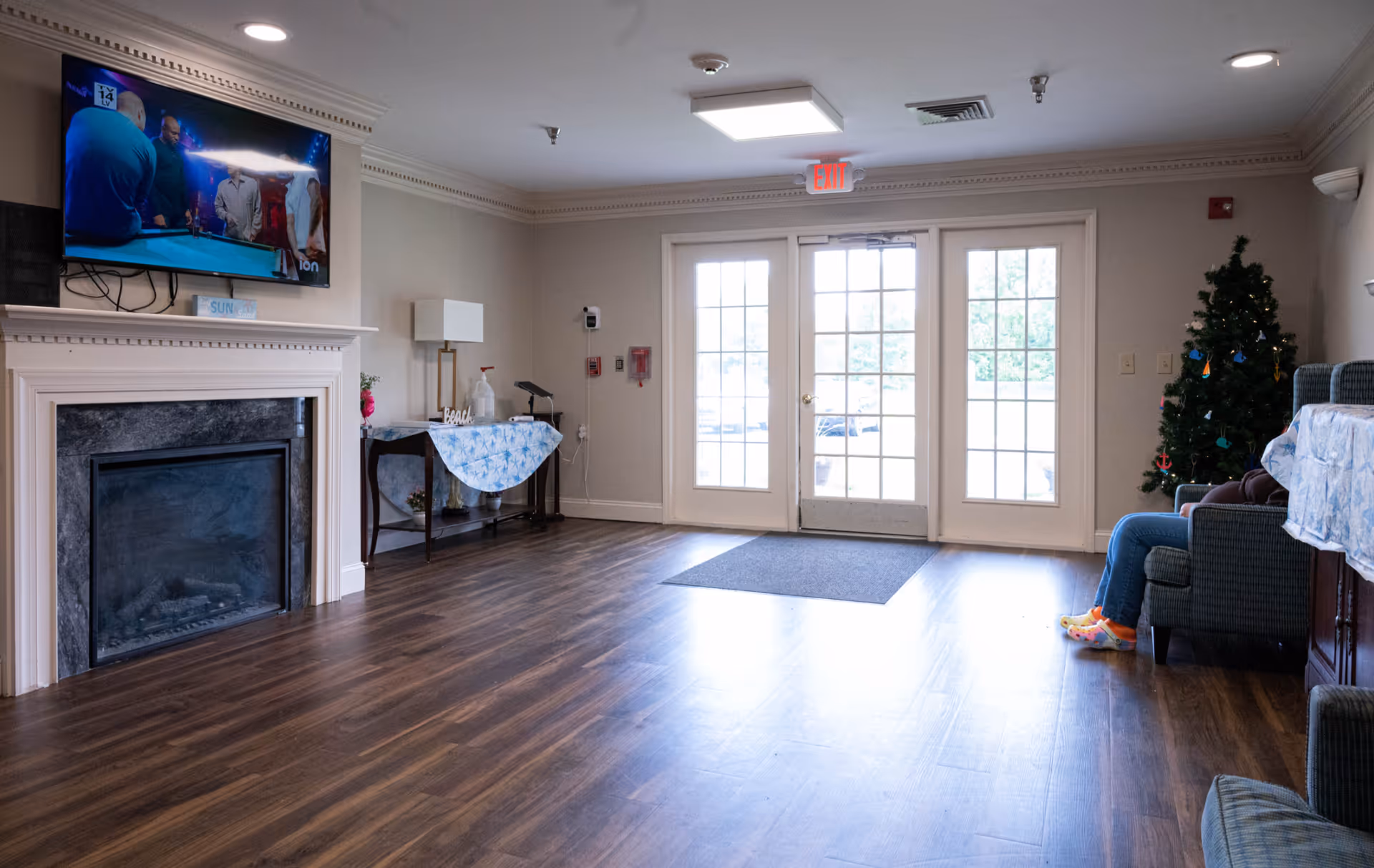 A cozy living room area in a senior living facility with a wall-mounted TV above a fireplace, a table with a lamp and decorative items, a Christmas tree, and a person sitting on a chair wearing colorful socks. The room has wooden flooring and glass double doors leading outside.