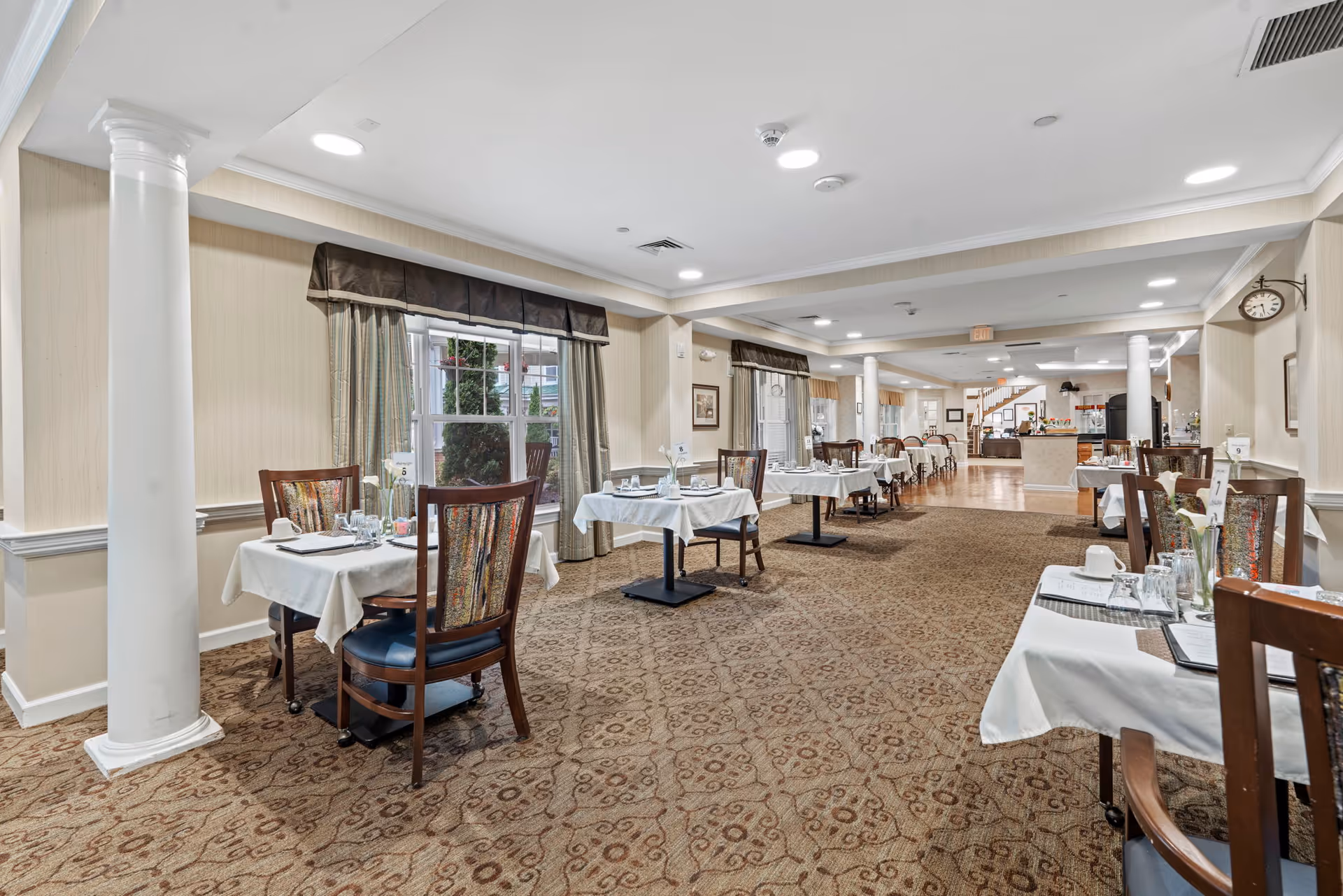 A spacious dining room with multiple tables covered in white tablecloths, each set with glasses, cups, and menus. The room features patterned carpet, beige walls, large windows with curtains, and white columns. There is a clock on the wall and a counter area visible in the background.