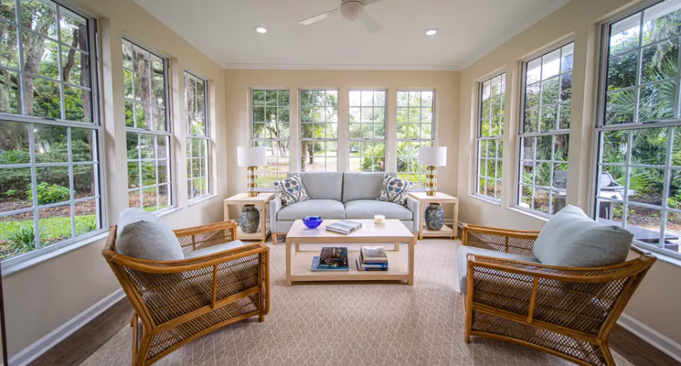 Bright sunroom-style living room with large windows, a central sofa, two wicker chairs, a coffee table, and side lamps.