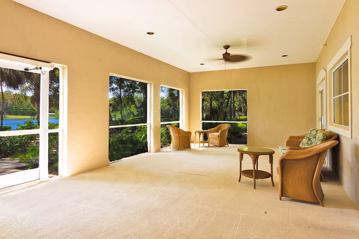 A spacious screened-in porch area with beige walls and ceiling, featuring wicker furniture including two armchairs with cushions, a small round table, and a loveseat with floral cushions. The porch overlooks a lush green outdoor area with trees and a body of water visible through the screened windows. A ceiling fan is mounted on the white ceiling.