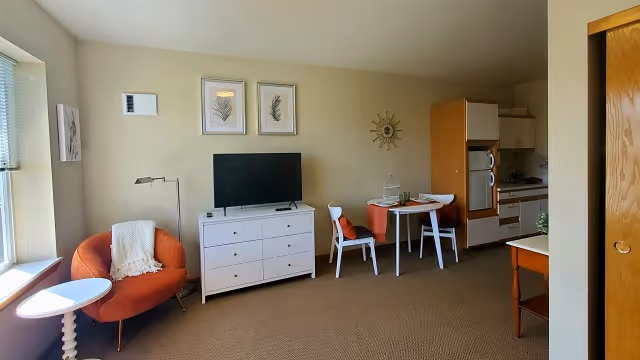 Studio-style living area with a TV on a white dresser, an orange armchair by a window, a small dining table, and a kitchenette to the right.
