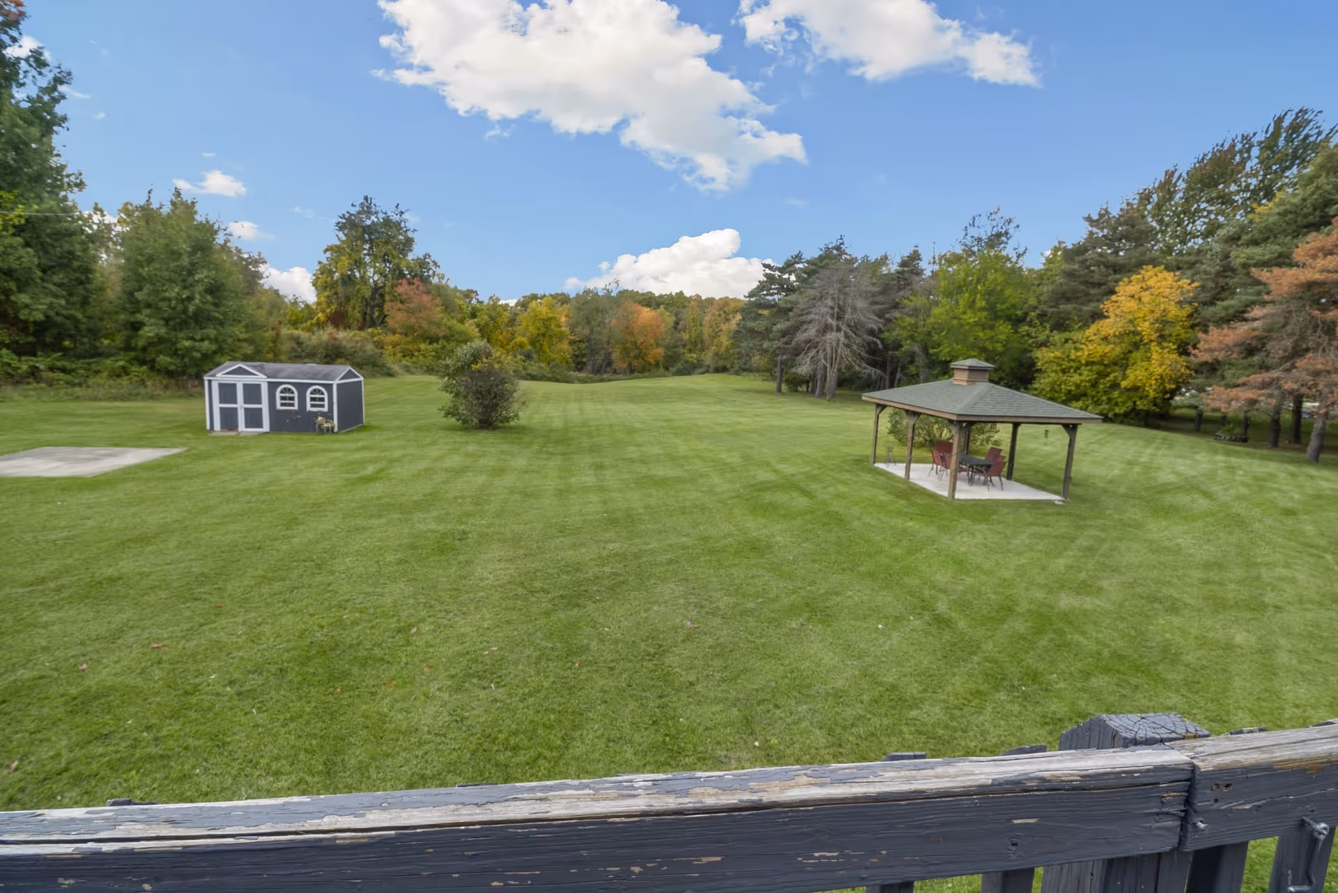 A large, well-maintained grassy outdoor area with a small shed on the left and a gazebo with chairs and a table on the right. The background features a variety of trees with some showing autumn colors under a partly cloudy blue sky.