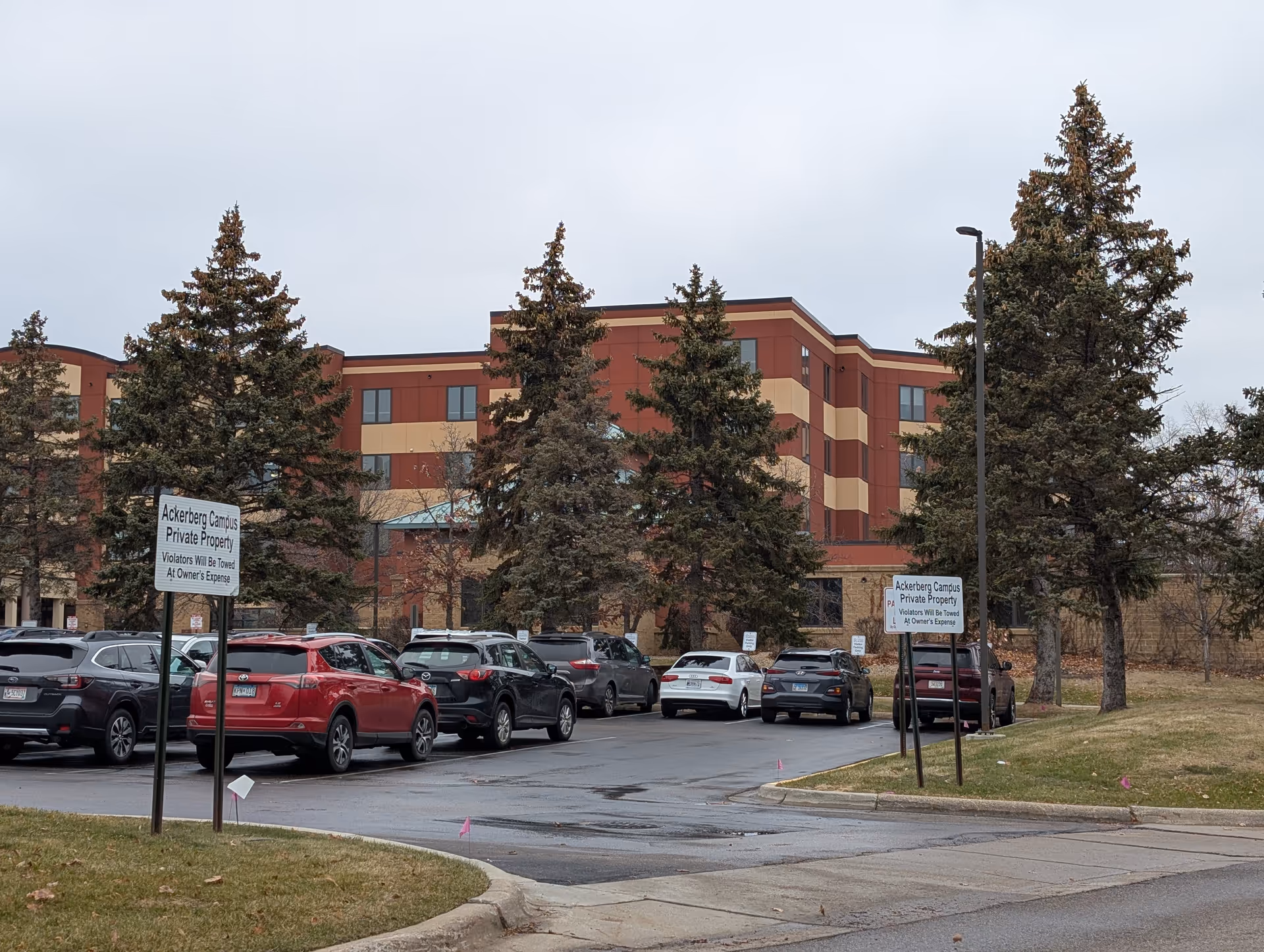 Exterior front of a multi-story assisted living building with a parking lot, several parked cars, and tall evergreen trees.