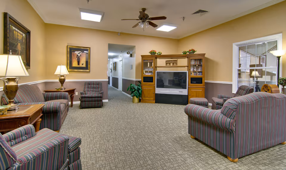 A cozy senior living facility common area with striped upholstered armchairs and sofas arranged around a wooden entertainment center holding a large TV. The room has beige walls with white trim, carpeted floor, ceiling fan with lights, table lamps, framed artwork, and a hallway leading to other rooms.