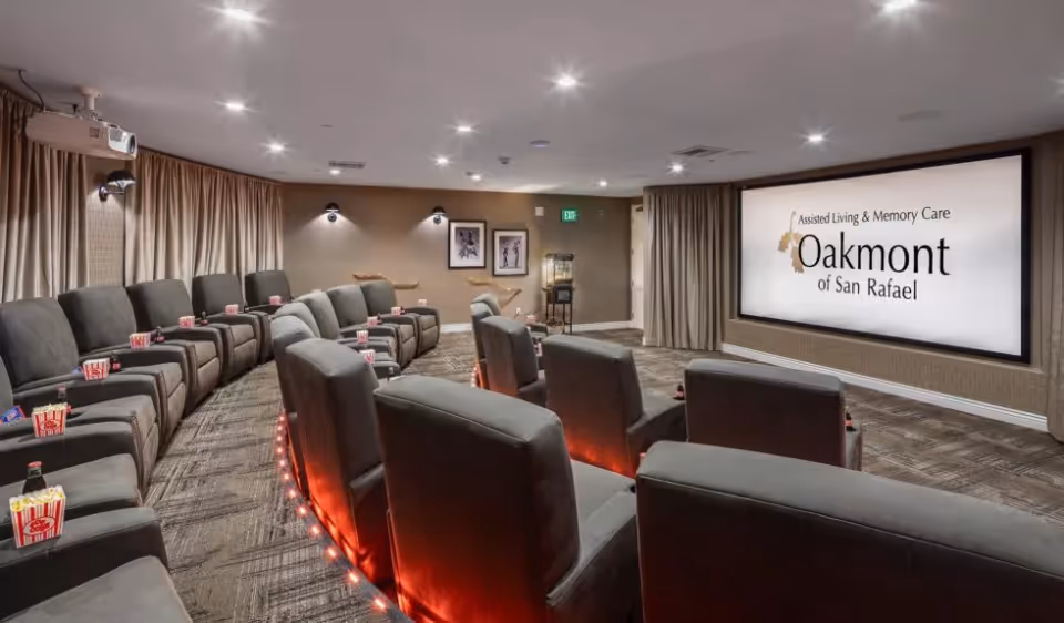 A cozy screening room with tiered reclining theater chairs, cupholders and a large projection screen showing 'Oakmont of San Rafael'.