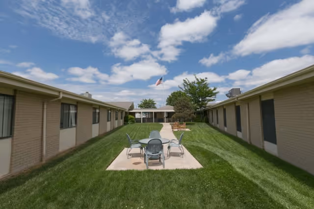 Grassy courtyard between two single-story brick building wings with outdoor tables and chairs and an American flag under a partly cloudy sky.