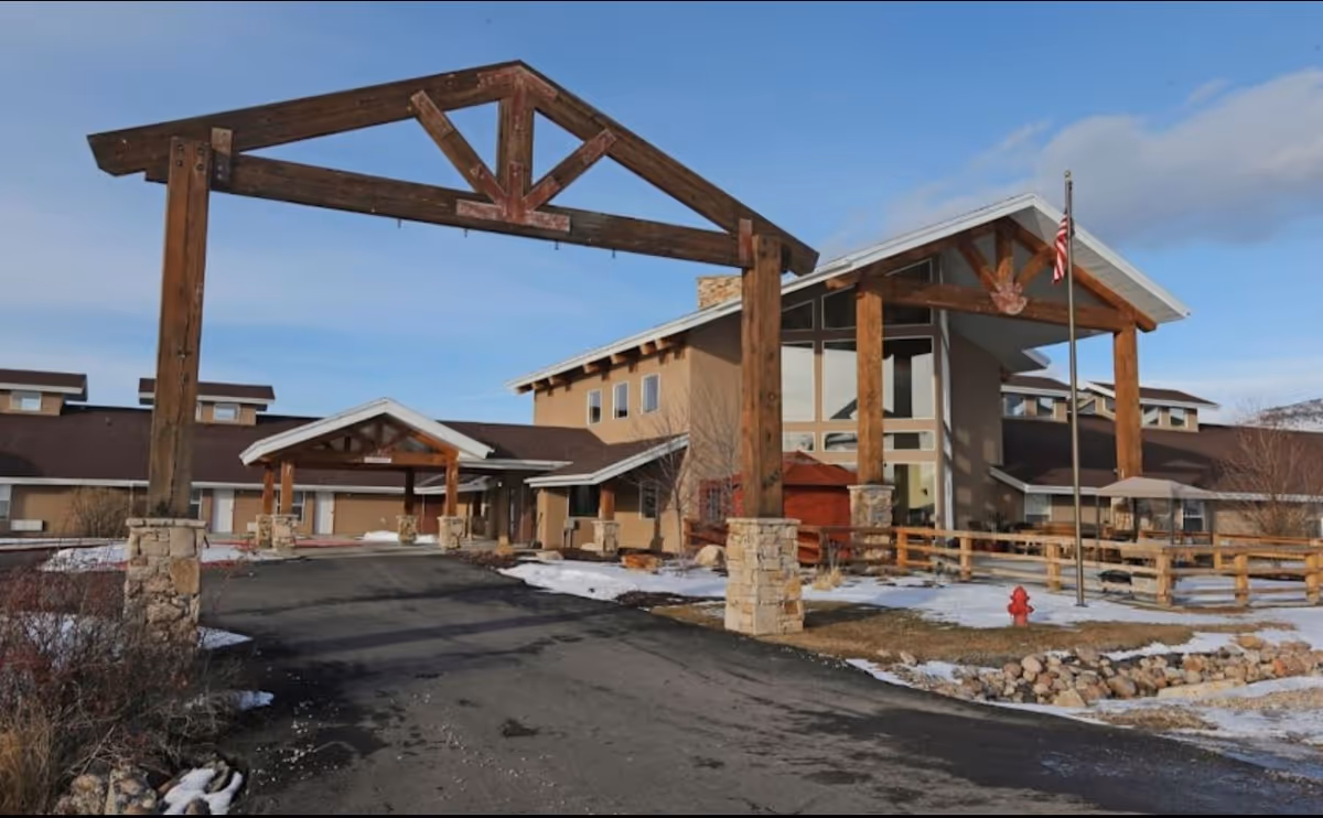 Front exterior of a large assisted living building with prominent wooden beams, a covered entrance, and light snow on the ground.