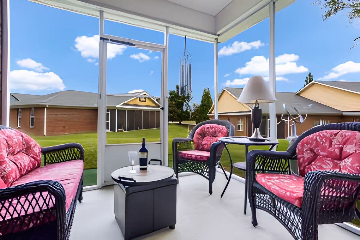 Screened-in porch with black wicker furniture featuring pink cushions, a small table with a bottle and glass, and a side table with a lamp. The porch overlooks a grassy area with buildings and trees under a blue sky with scattered clouds.