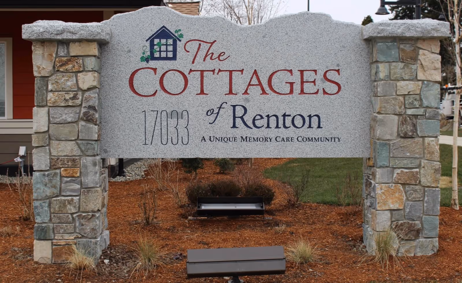Stone sign for The Cottages of Renton, a unique memory care community, with the address 17033 displayed. The sign is supported by two stone pillars and is set in a landscaped area with mulch and some plants.