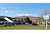 Front exterior of Holbrook Adult Home: a single-story building with a parking lot, several cars, a grassy lawn, and a roadside sign, set against distant hills.