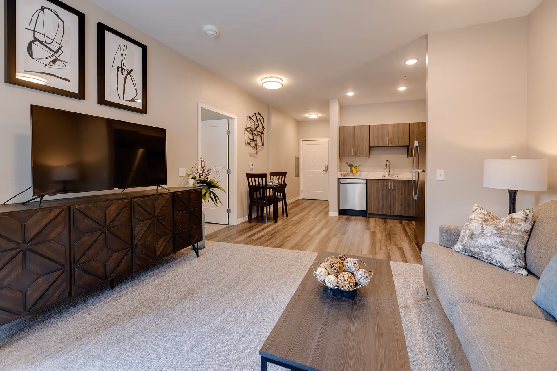 Open-concept living room and kitchenette with a sofa, coffee table, TV on a wood console, dining table, and modern kitchen cabinets.