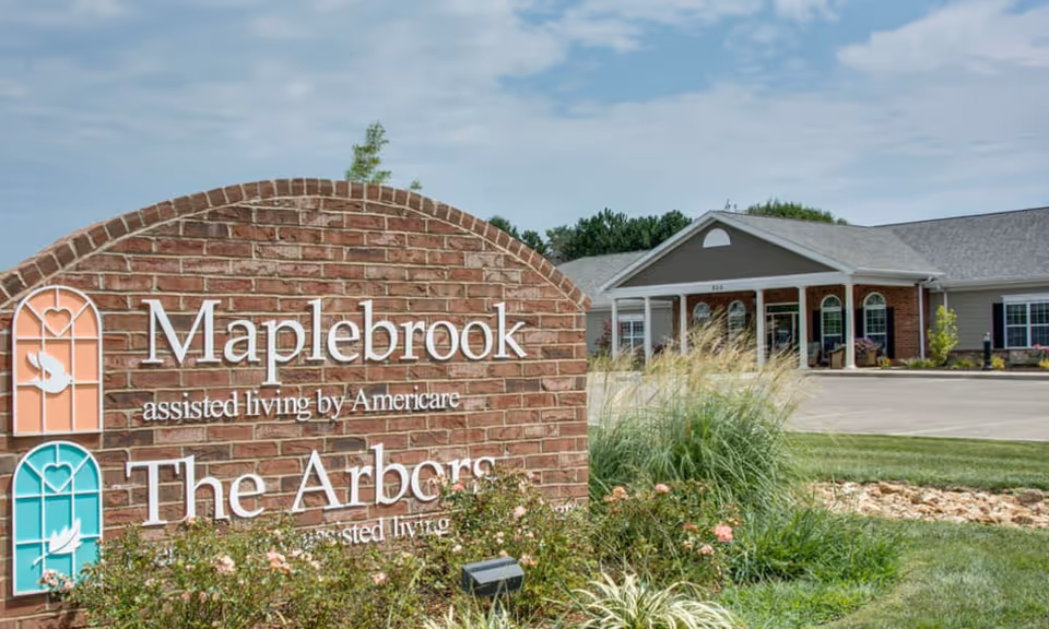 A brick entry sign for Maplebrook and The Arbors in front of a single-story senior living community building and landscaped grounds.