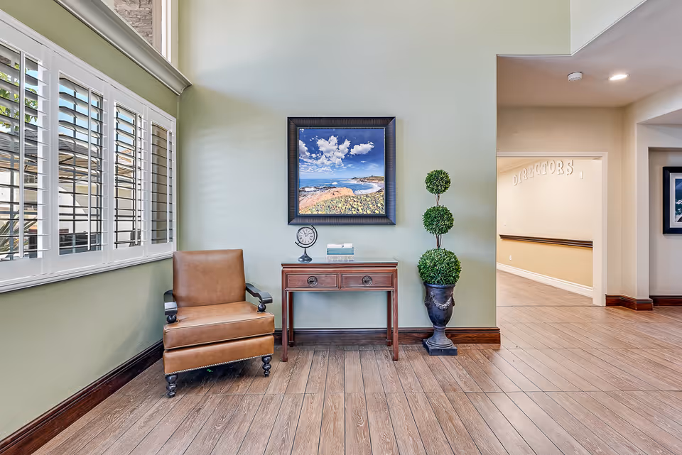 A cozy corner in a senior living facility with a brown leather armchair, a small wooden table holding a clock and books, a decorative topiary plant in a black vase, and a framed landscape painting on a light green wall. Large windows with white shutters allow natural light to fill the space, and a hallway labeled 'DIRECTORS' is visible in the background.