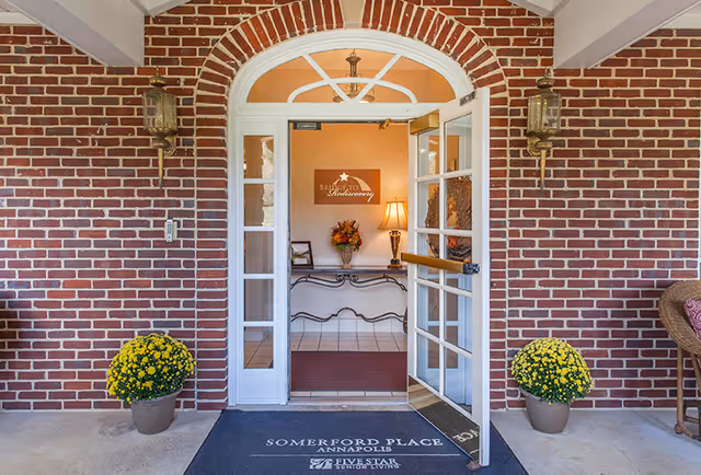 Brick entrance with open glass double doors leading into a warmly lit lobby, flanked by potted yellow flowers.