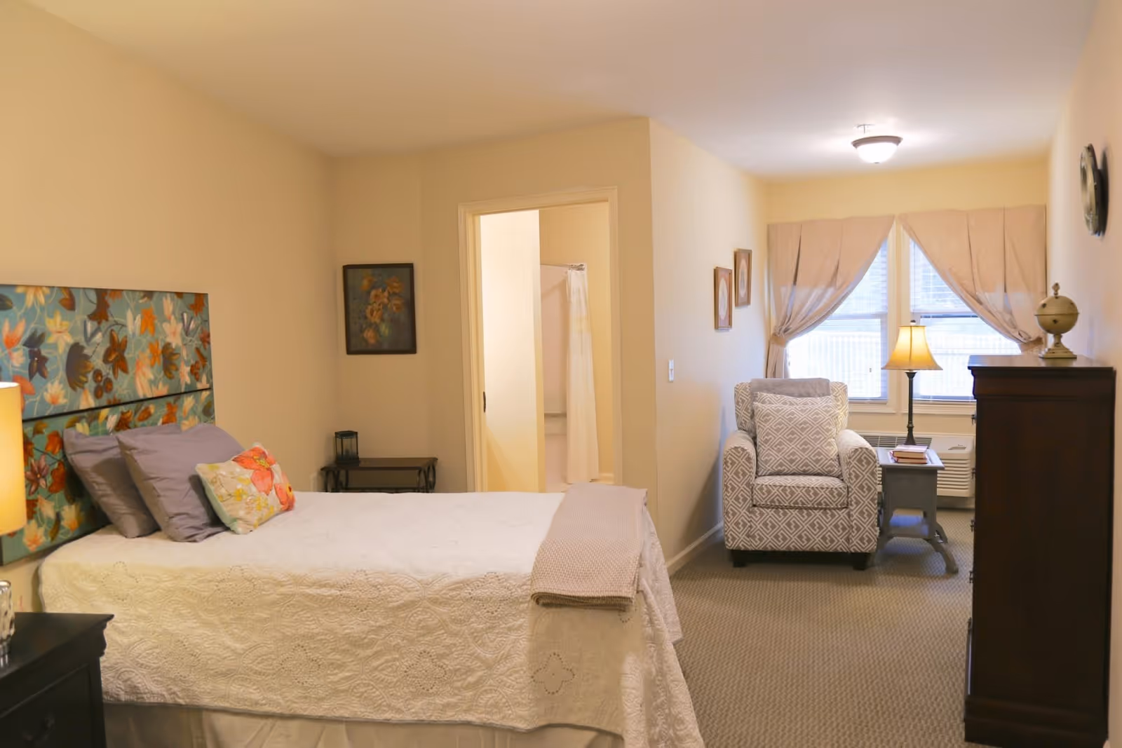 A cozy bedroom in a senior care facility featuring a bed with a floral headboard, multiple pillows, and a white quilt. There is a small table with a lamp and a chair near a window with beige curtains. The room has beige walls, carpeted floor, and a doorway leading to a bathroom with a shower curtain visible.